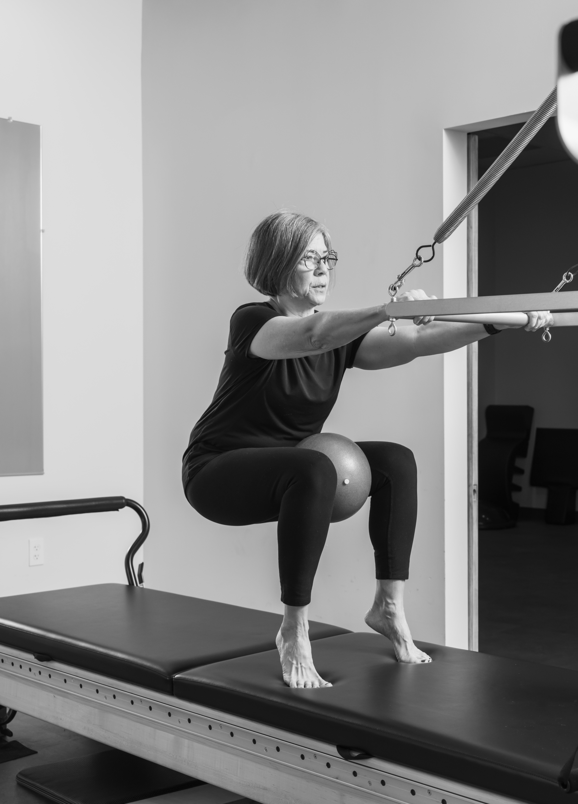 Woman performing a squat exercise on a Pilates reformer, holding the spring-loaded bar with a small ball squeezed between her knees.