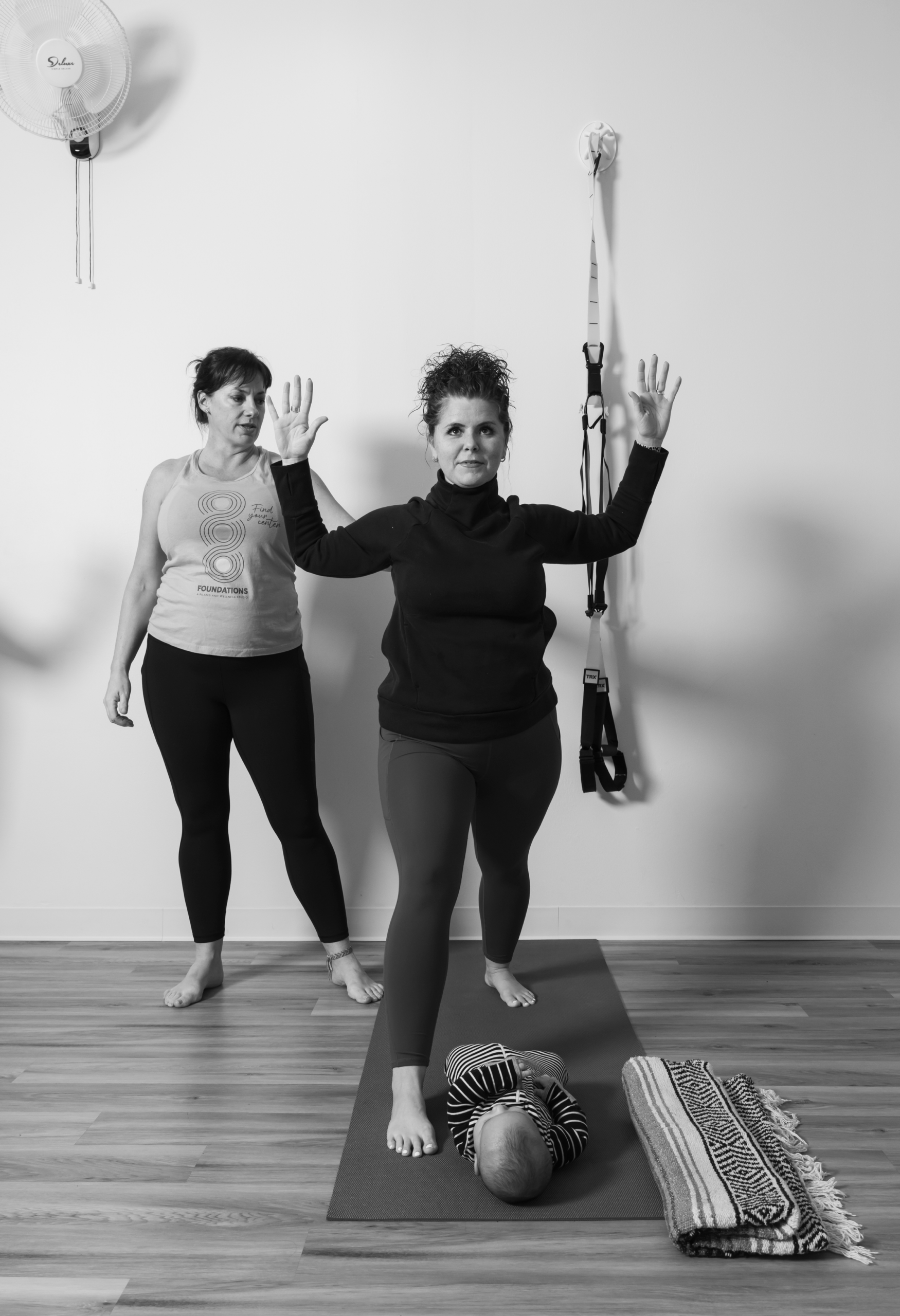 Foundations Studio postpartum pilates instructor guiding a mother through an arm balance exercise, with her baby resting on the yoga mat at her feet